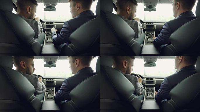 两名男子汽车经销商和顾客坐在漂亮的汽车前排座位上讨论汽车型号的后视图。皮革座椅和电子板是可见的。