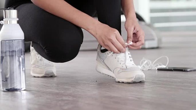 女跑步者的脚特写镜头准备用瓶装水、耳机和电话系跑鞋，以激发室内健身房运动跑道上有氧运动训练的音乐动机