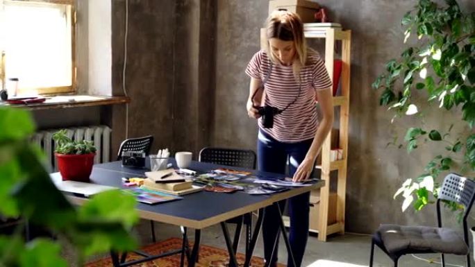 年轻的创意女孩正在用彩色照片平铺，然后用相机拍摄。女人站在现代餐桌旁，穿着便服。