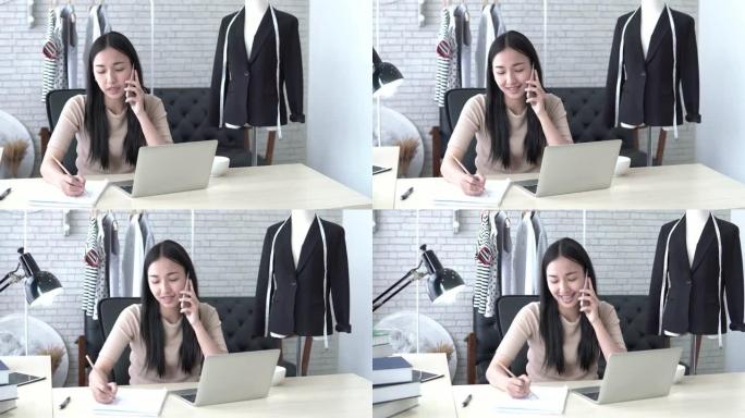 4k分辨率的亚洲女设计师坐在桌面上，在她的时装工作室里用新衣服的草图在电话里聊天