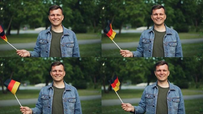 慢动作肖像欢快的德国游客挥舞着德国的官方旗帜，微笑着看着镜头。背景是美丽的公园，温暖的夏日。