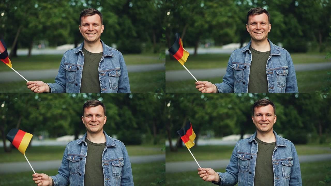 慢动作肖像欢快的德国游客挥舞着德国的官方旗帜，微笑着看着镜头。背景是美丽的公园，温暖的夏日。