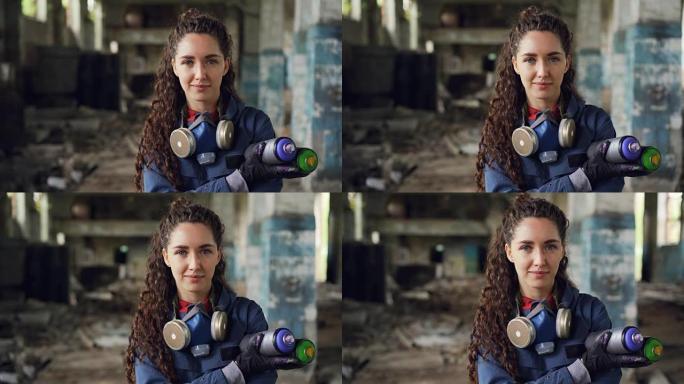 微笑女孩涂鸦艺术家的特写肖像站在废弃的建筑里，拿着喷漆，看着相机。女人有防护口罩和手套。