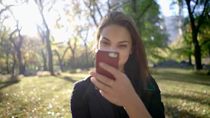 年轻漂亮的黑发女人在阳光明媚的日子里使用智能手机。秋天公园，落叶和树木。