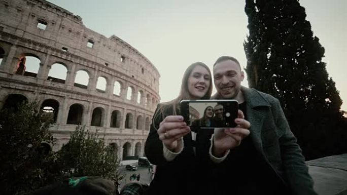 年轻美丽的夫妇坐在意大利罗马的罗马斗兽场附近，一起在智能手机上自拍