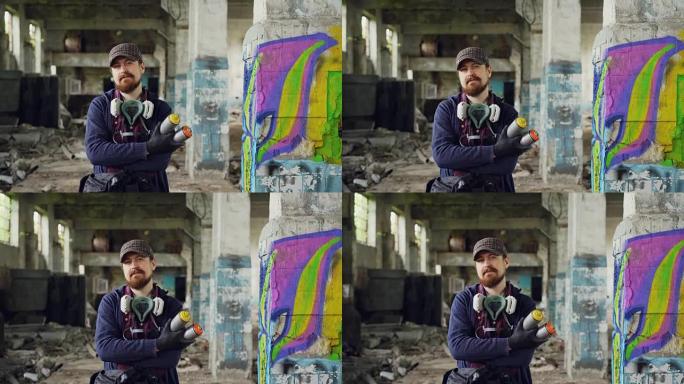 大胡子的盖伊涂鸦画家的肖像站在古老的空荡荡的建筑物中，背景是涂鸦，并拿着喷漆。男人戴着防毒面具，戴着