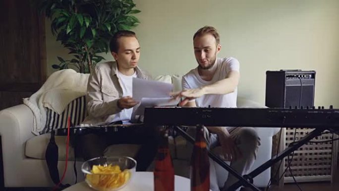 吉他手和键盘手正在排练休息时看着乐谱并讨论歌词。电子乐器，饮料和小吃是可见的。