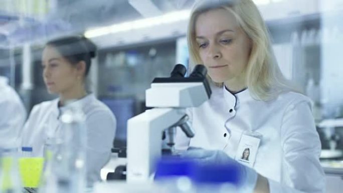 女性研究科学家将滑倒给她的同事，后者在显微镜下观察样品。他们在一个大的研究中心/实验室工作。