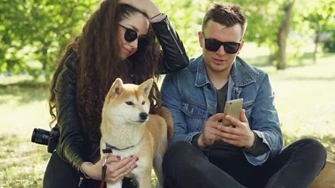 年轻女子抚摸着她的狗坐在草地上，而她的男朋友正在显示她的智能手机屏幕，人们在说笑。公园里的周末概念。