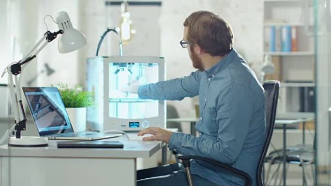年轻的大胡子开发人员使用3D打印机进行建模。他坐在技术现代化的办公室里的办公桌前。