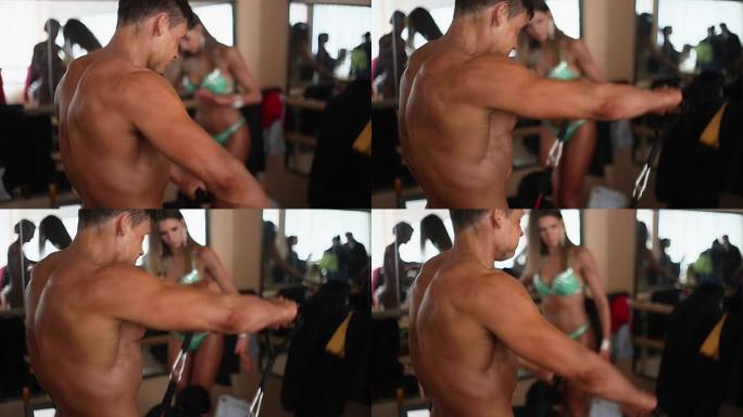 侧面，后视图的男性健美运动员，裸露的躯干做阻力带运动。背景中穿着比基尼的女人