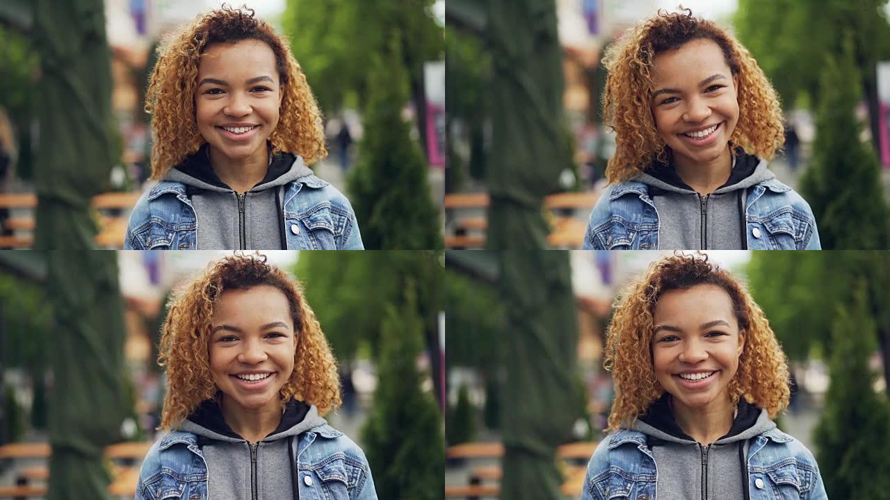 夏天，迷人的非洲裔美国女孩看着相机，微笑，大笑，抚摸着她的头发，站在美丽的绿色公园。