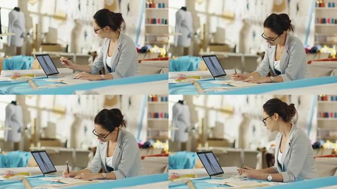 美丽的女性时装设计师坐在办公桌前看着笔记本电脑上的草图，做笔记。她的工作室阳光明媚，色彩丰富的面料和