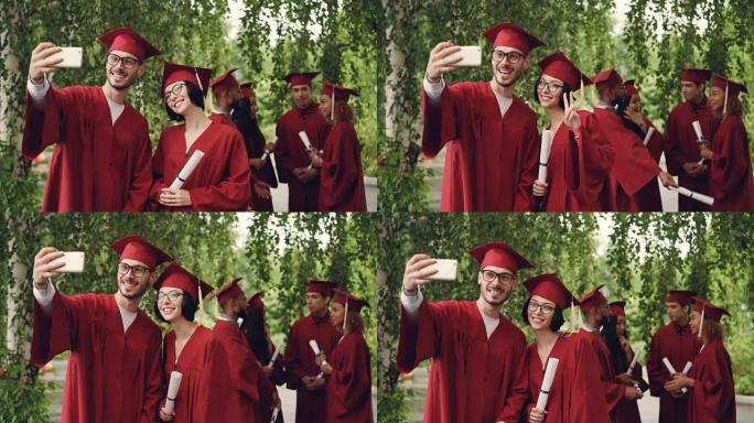 开朗的一对即将毕业的学生正在使用智能手机进行自拍照，年轻的男人和女人拿着文凭，看着智能手机摄像头并微