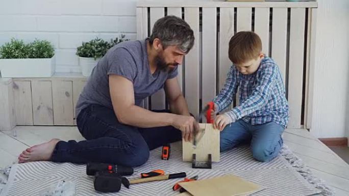 乐于助人的儿子正在用手锯锯木头，而他的父亲正在用量筒测量切口并与孩子交谈。建筑、家庭和童年的概念。