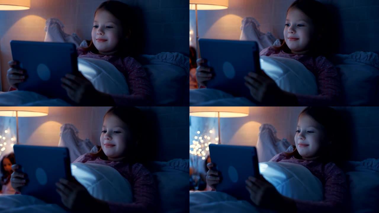 晚上快乐的小女孩躺在床上，在平板电脑上看东西。她的床灯亮着。