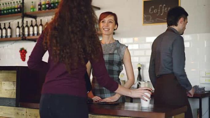 nice modern cafe的礼貌微笑的员工正在出售外卖饮料，而快乐的顾客则使用智能手机付款。电