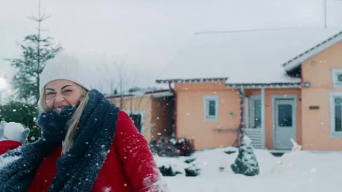 快乐的年轻微笑的女人扔雪球，被一个打中。穿着红色外套的女孩和她的伴侣一起享受冬天。田园诗般的房子后院