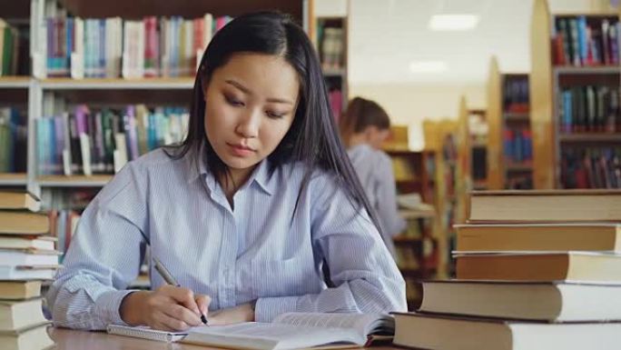 年轻漂亮的亚洲女学生坐在大图书馆里被成堆的书包围的桌子旁，重写课本上的文字准备考试