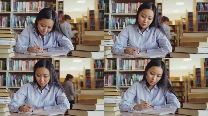 年轻漂亮的亚洲女学生坐在大图书馆里被成堆的书包围的桌子旁，重写课本上的文字准备考试