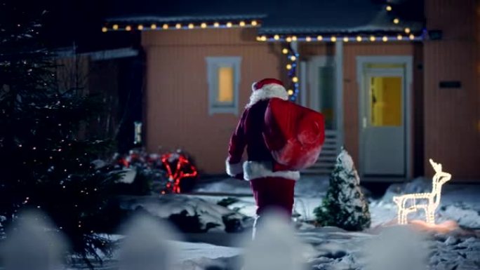 地道的圣诞老人肩上扛着红色的包，走进装饰着灯光和花环的田园诗般的房子的前院。圣诞老人带来礼物和礼物。