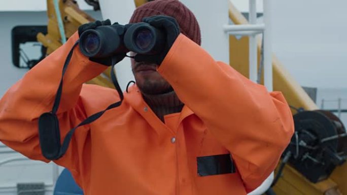 穿着防护外套的商业渔船船长透过双筒望远镜看