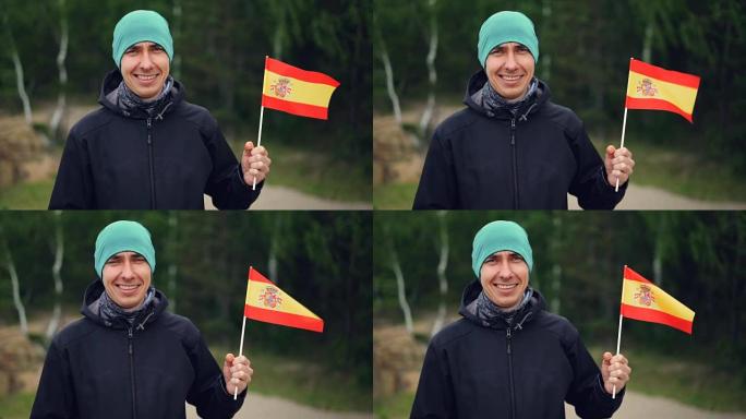 微笑的年轻人拿着西班牙国旗，看着站在绿色森林附近的相机的慢动作肖像。人和民族概念。