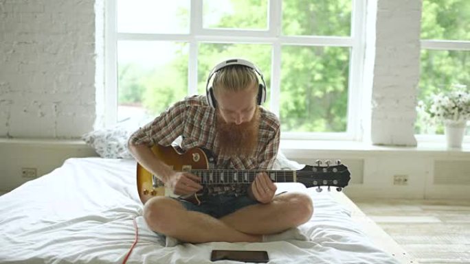戴着耳机的有胡子的男人坐在床上学习在现代卧室用平板电脑弹吉他