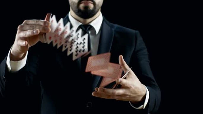 穿着黑色西装表演纸牌把戏的熟练魔术师的特写镜头。在空中投掷和捕捉纸牌。背景是黑色的。慢动作。