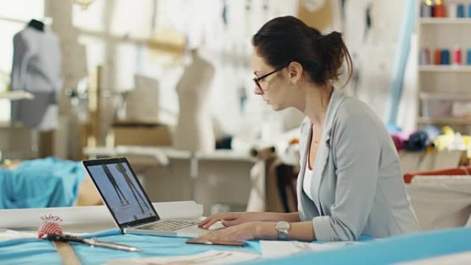 美丽的女性时装设计师坐在她的桌子上，在笔记本电脑上工作，看着图纸和草图。她的工作室阳光明媚，色彩丰富
