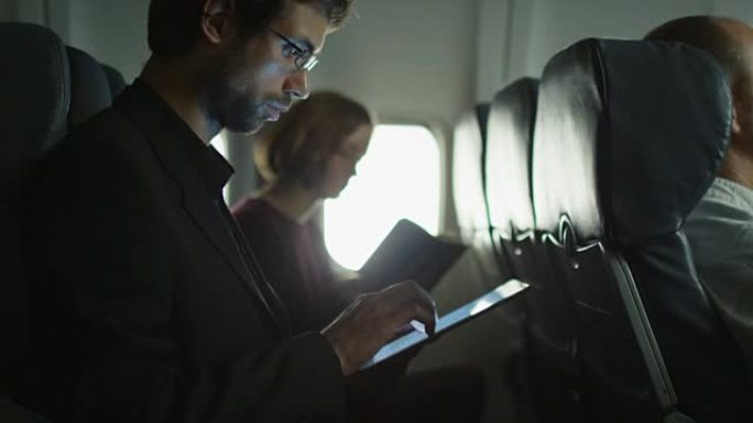 年轻人在飞机上使用平板电脑，一个女人在窗户旁边的背景中阅读。