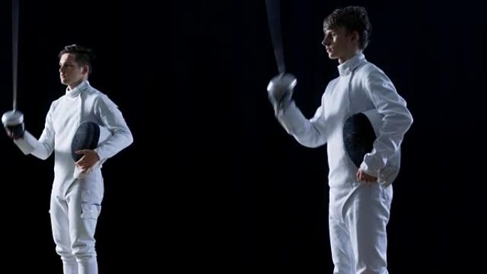 两个年轻的击剑手互相问候观众，戴上面具，用箔纸开始剑术比赛。在孤立的黑色背景上拍摄。