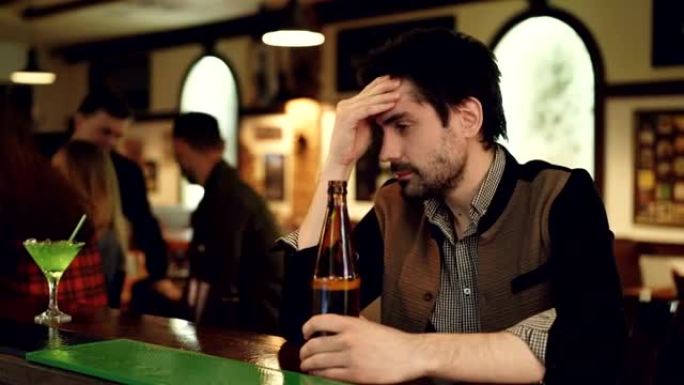 沮丧的未刮胡子的家伙正坐在酒吧柜台旁，手里拿着一瓶啤酒，叹息着，绝望地抚摸着他的脸。他看着年轻人玩得