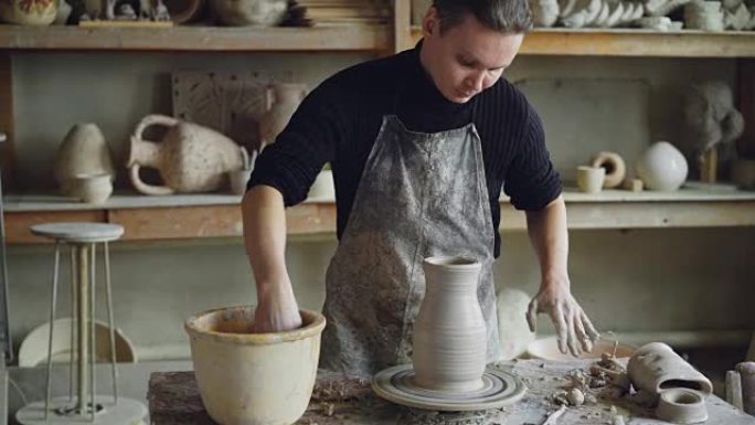 一名英俊的年轻人正在陶艺室工作，他正在纺车上用陶土塑造陶瓷花瓶。专业就是专注于工作。
