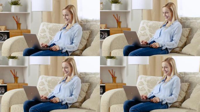 一个微笑的美女坐在客厅的沙发上的长镜头。她在笔记本电脑上工作，笔记本电脑放在腿上。