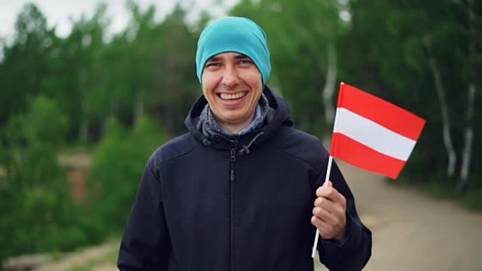 快乐的游客慢动作肖像，举着奥地利国旗，看着相机，站在异国绿色森林附近，微笑着。
