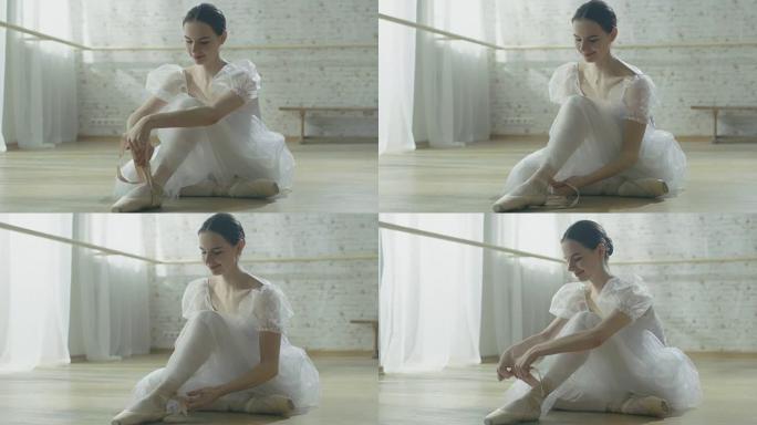 年轻漂亮的芭蕾舞演员穿着芭蕾舞短裙坐在木地板上，系着她的脚尖鞋。慢动作。