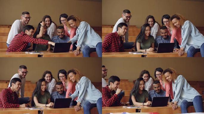 开朗的男人和女人正在用笔记本电脑看着并指向屏幕，坐在大学演讲厅的桌子上聊天和大笑。学生和技术概念。