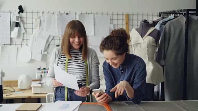 创意裁缝在nice studio工作时使用智能手机并查看服装草图。背景中的假人、缝纫机和女装。