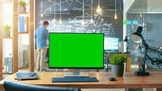 带有模拟绿屏监视器的个人计算机站在办公桌上，背景是白天在创意办公室工作的人。
