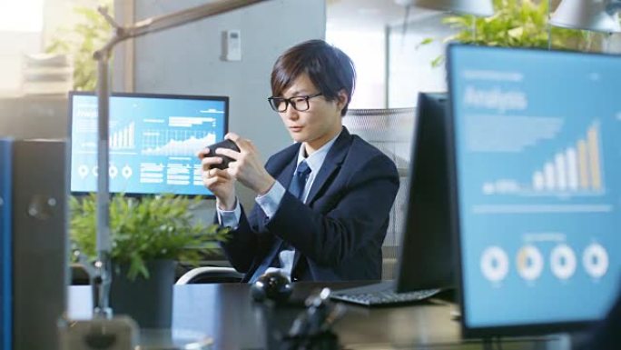 在办公室里，东亚商人在智能手机上玩电子游戏。微笑的帅哥坐在办公桌前。