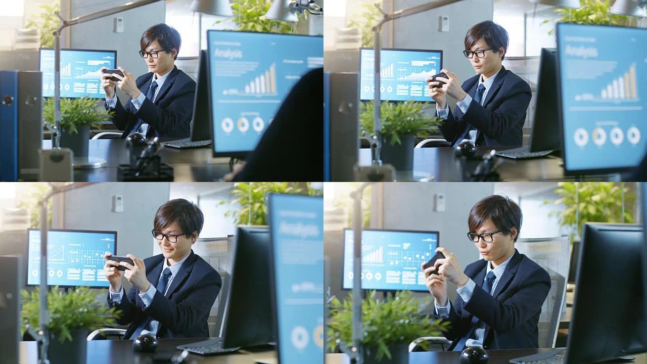 在办公室里，东亚商人在智能手机上玩电子游戏。微笑的帅哥坐在办公桌前。