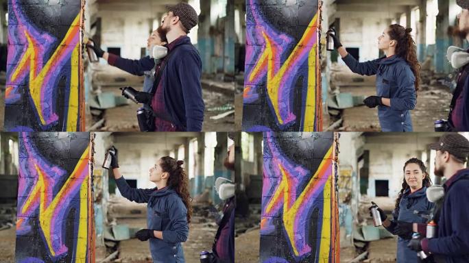 开朗的年轻女子业余涂鸦艺术家正在学习用熟练的大胡子画家喷漆，同时在空仓库里装饰旧柱子。