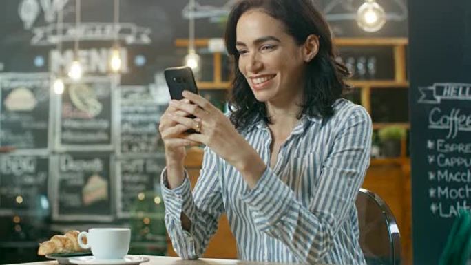 美丽的女人坐在咖啡馆里使用智能手机。她在给朋友或爱人发消息时微笑。在她端着饮料和羊角面包的桌上杯子上