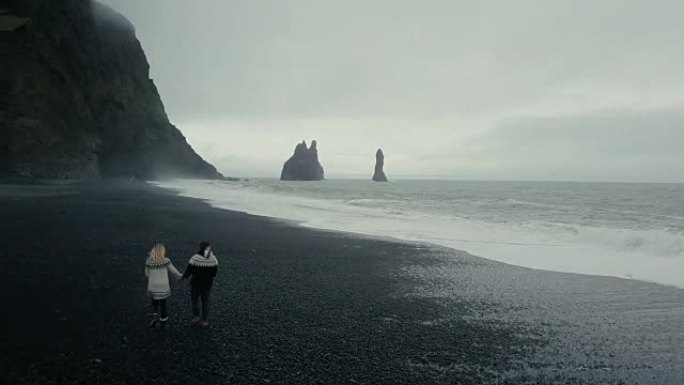 在冰岛巨魔脚趾悬崖附近的黑色火山海滩上散步的年轻夫妇的鸟瞰图