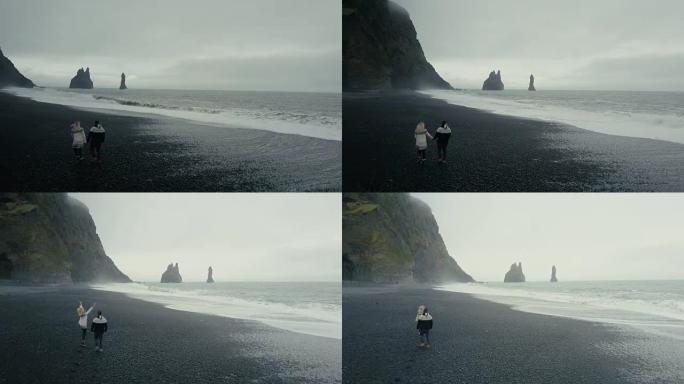 在冰岛巨魔脚趾悬崖附近的黑色火山海滩上散步的年轻夫妇的鸟瞰图