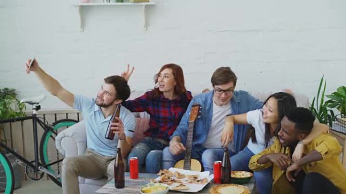 快乐的学生朋友在智能手机上自拍，在室内共享住宿中与啤酒和比萨饼聚会时摆姿势