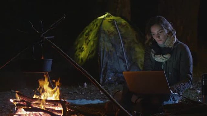 年轻的黑发女子晚上在篝火旁使用笔记本电脑。