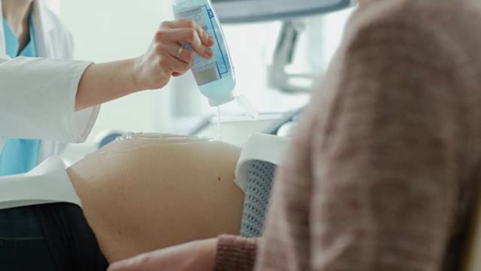在医院里，女产科医生将凝胶涂在一位孕妇的肚子上，准备做超声波筛查。父亲勤奋地支持他怀孕的妻子。幸福的
