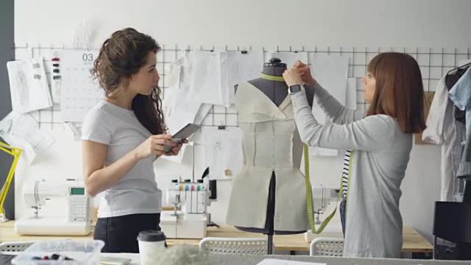 服装设计师企业家在漂亮的现代工作室工作时正在测量人体模型的零件。年轻迷人的女人正在使用平板电脑输入测